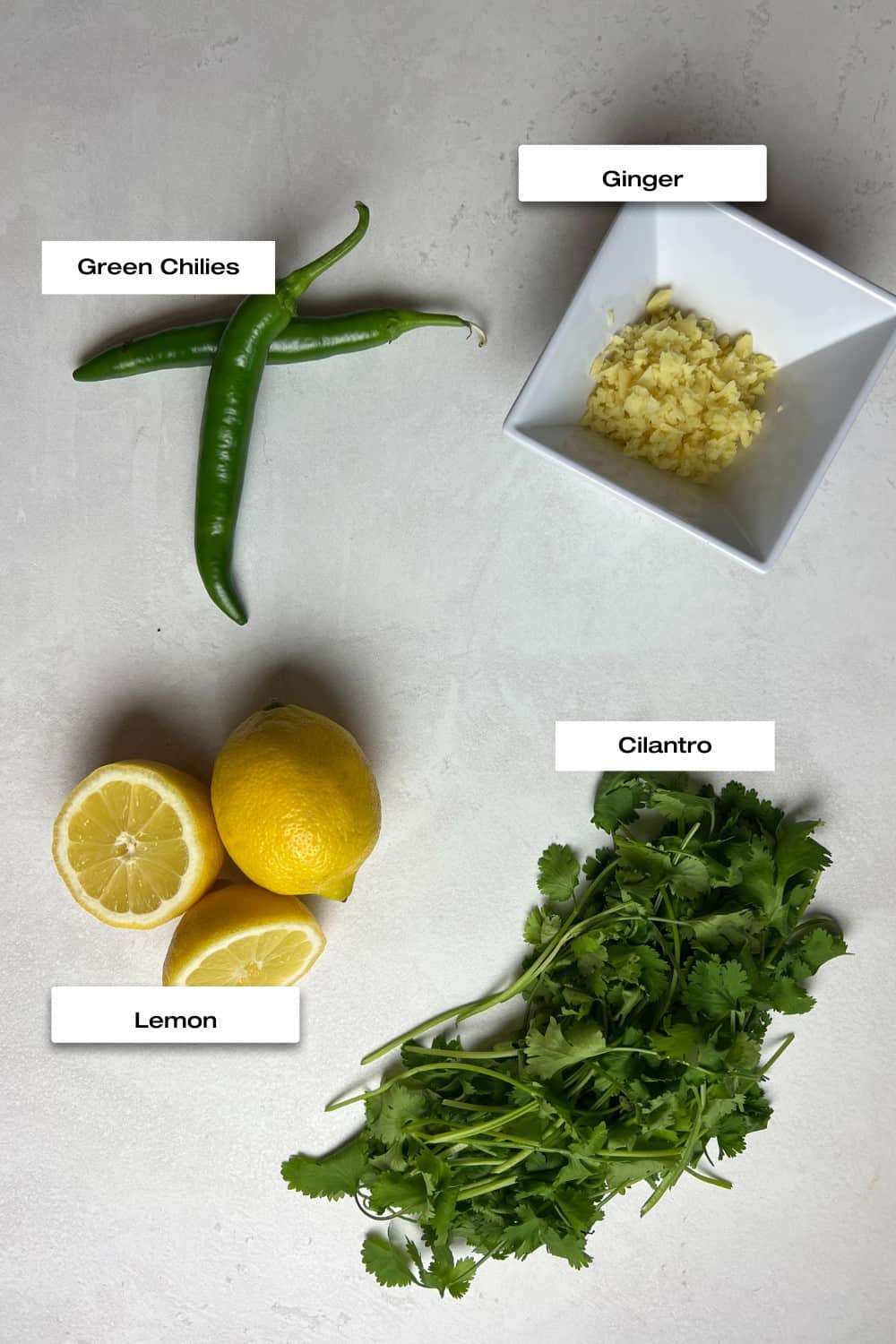 White countertop with green chilies, lemons, cilantro and crushed garlic.