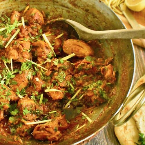 Cooked chicken karahi in a stainless steel karahi with a wooden spoon and naan and lemons in the background.