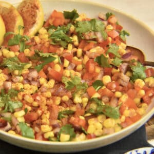 yellow corn, red onion, multi colored bell peppers and green cilantro mixed in a white bowl with lemon wedges.