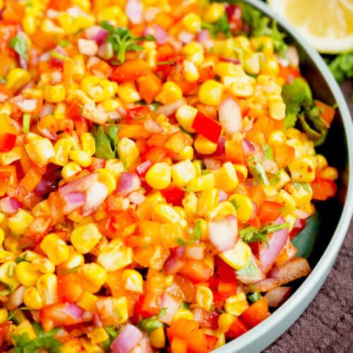 yellow corn, red onion, multi colored bell peppers and green cilantro mixed in a white bowl with lemon wedges.