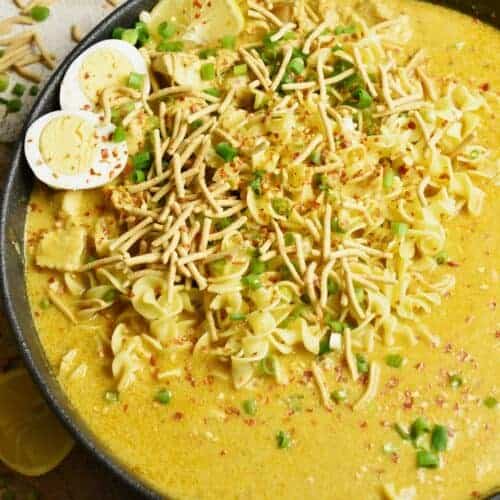 A large black bowl filled with a yellow coconut curry, egg noodles, crunchy noodles, half a boiled egg, quartered lemons, chopped green onions and red pepper flakes rests on a kitchen counter with a dark brown napkin and scattered crunchy noodles.