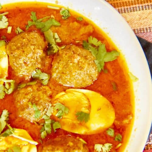 A white bowl filled with meatballs and halved hard boiled eggs floating in a red curry garnished with cilantro is resting on a blue napkin on a striped table cover.