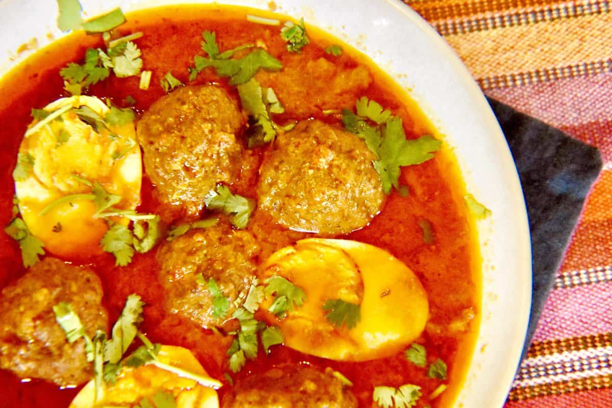 A white bowl filled with meatballs and halved hard boiled eggs floating in a red curry garnished with cilantro is resting on a striped table cover.