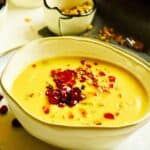 A light grey bowl resting on a light grey plate is filled with a yellow sweet potato lentil soup and garnished with a red chili oil, red pomegranate seeds and green toasted pistachios.