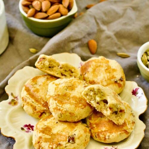 A fine china plate filled with Nan Khatai is resting on a grey napkin with bowls of almonds and cardamom in the background.