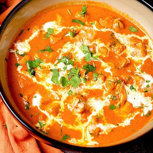 Cooked butter chicken curry, garnished with a swirl of cream and cilantro is being served in a light brown bowl with a dark brown rim.