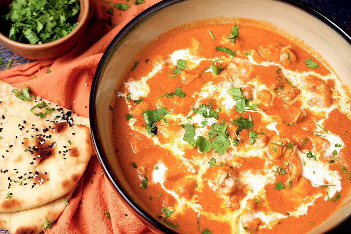 cooked butter chicken curry garnished with cream and cilantro is served with a side of naan.