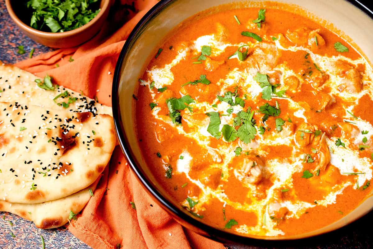 Cooked butter chicken curry is in a light brown bowl with naan and cilantro on the side.