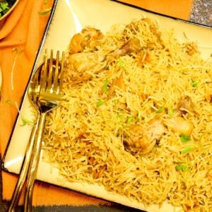 A pakistani chicken pulao ,plated in a beige platter, with a golden spoon and fork on the side, is resting on an orange napkin.