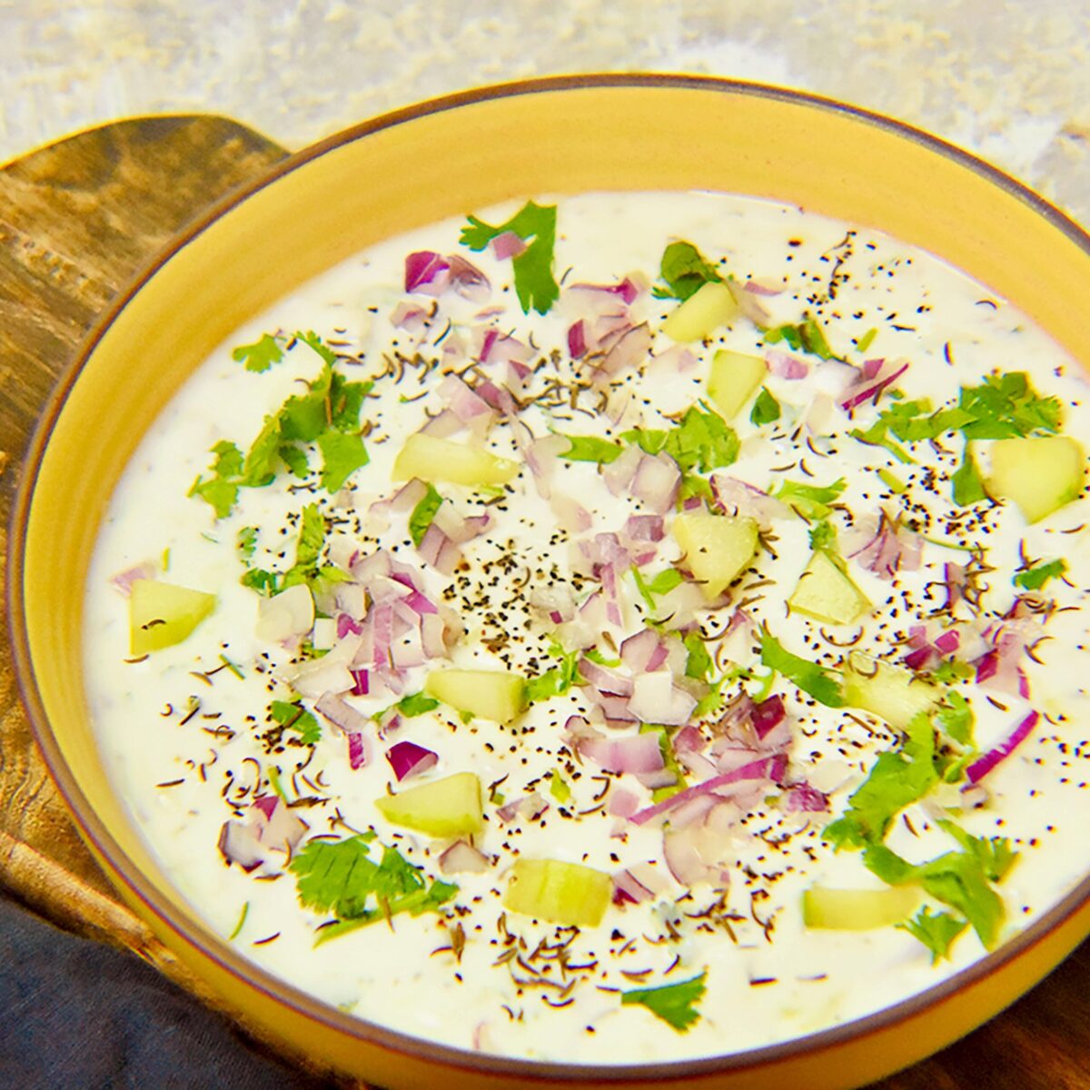 Cucumber raita in a yellow bowl sitting on a wooden board with a dark blue napkin on a light grey kitchen counter.
