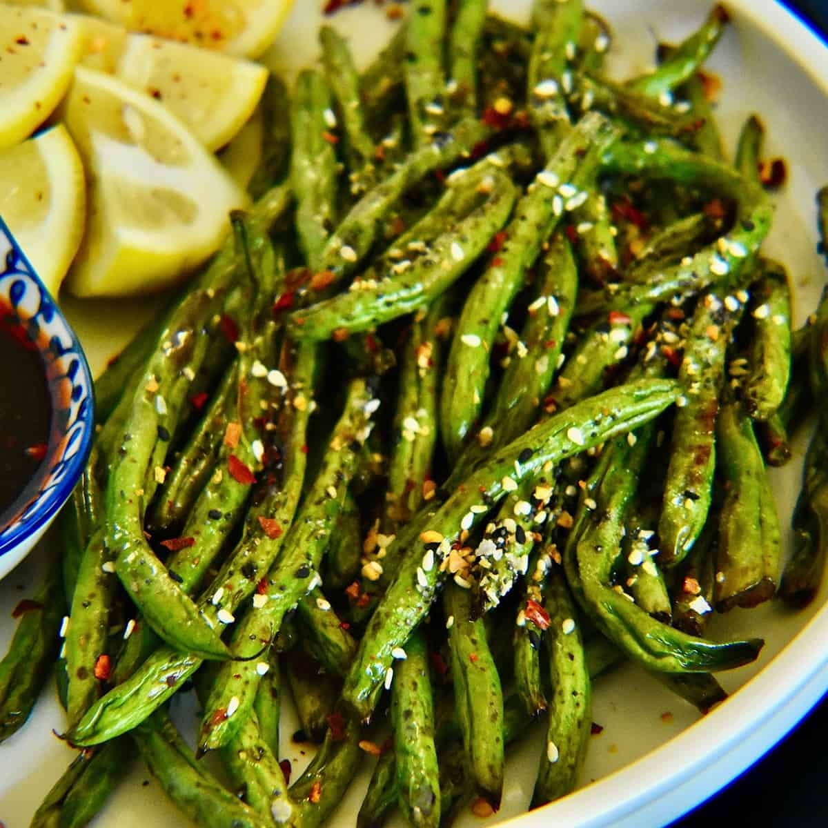 green beans in the air fryer are served on a white plate and served with lemon wedges and a sweet and spicy chili sauce.