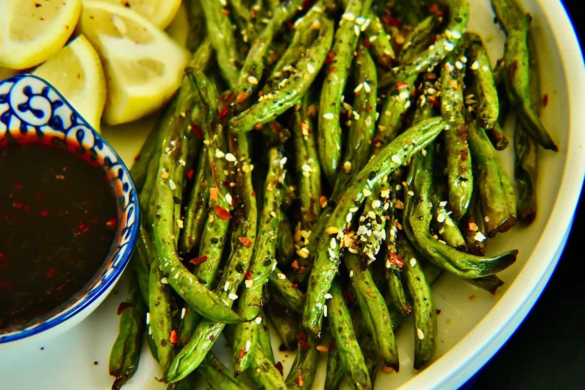 green beans in the air fryer are served on a white plate and served with lemon wedges and a sweet and spicy chili sauce.