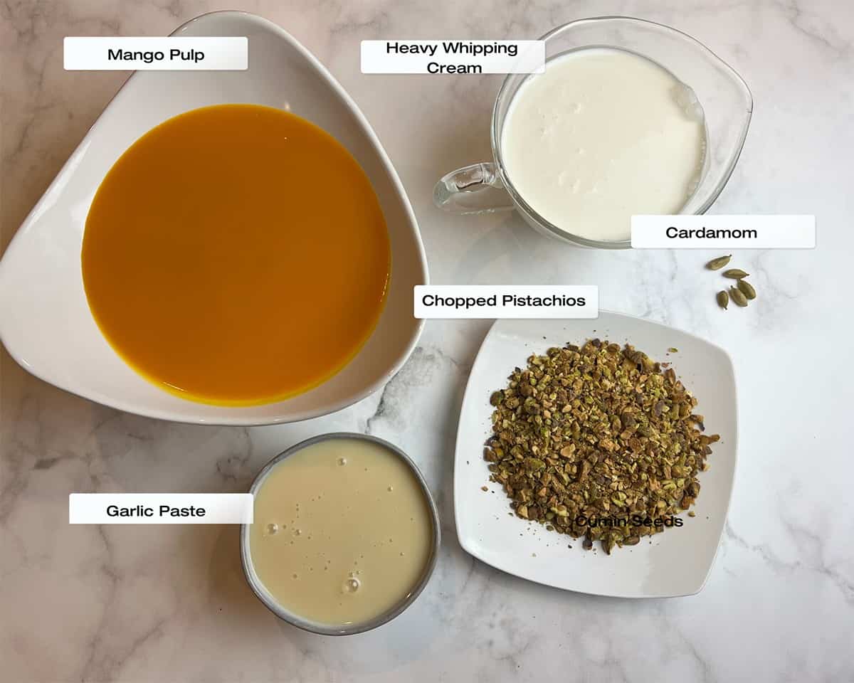 white marble counter-top with assorted bowls filled with mango pulp, condensed milk, heavy whipping cream, chopped pistachios and cardamom to make mango kulfi.