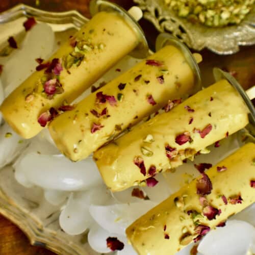 mango kulfi garnished with roses and pistachios on a bed of ice in a silver tray.
