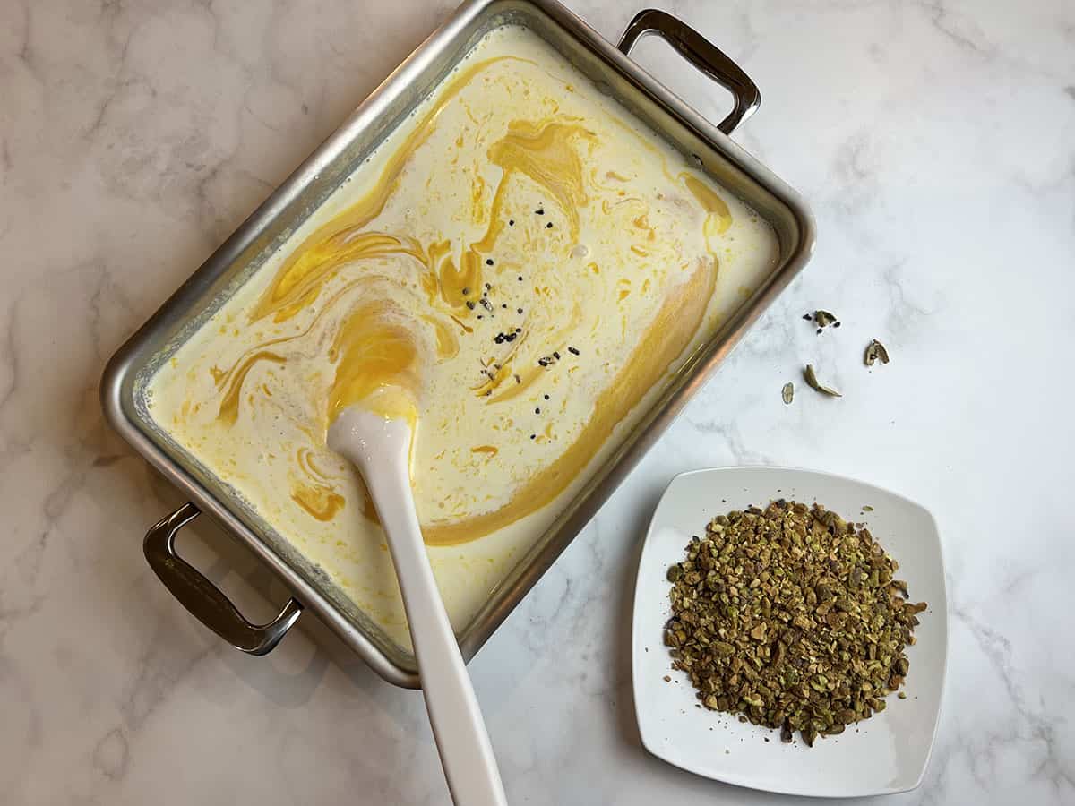 A white counter-top with a rectangular metal pan filled with a mixture for mango kulfi and a spoon to stir it, with a bowl of crushed pistachios on the side.