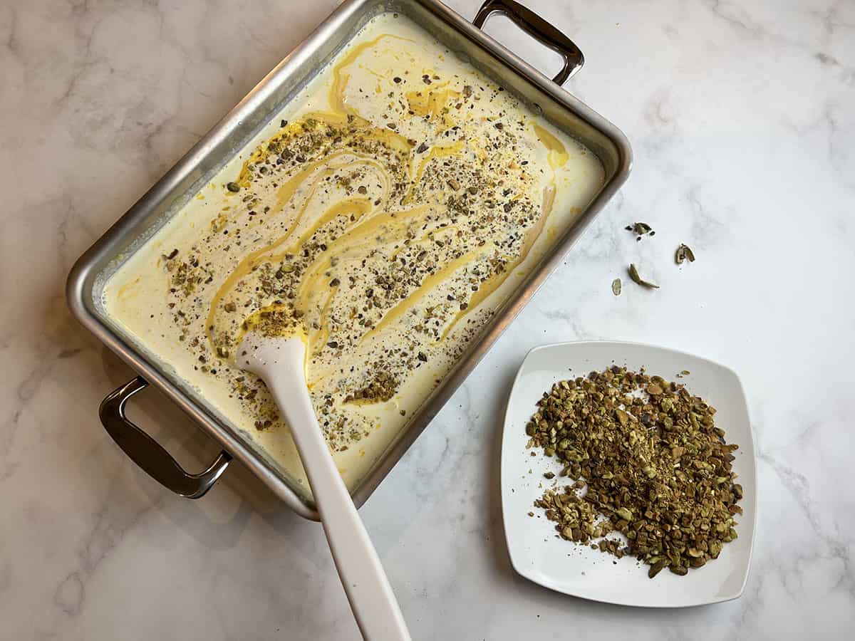 A white counter-top with a rectangular metal tray filled with a mango kulfi mixture and chopped pistachios being stirred.