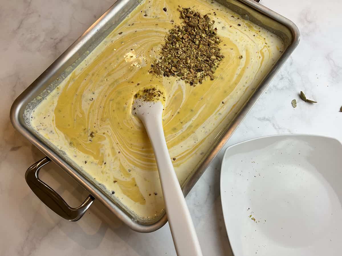 A white counter-top with a rectangular metal tray filled with a mango kulfi mixture and chopped pistachios being stirred.