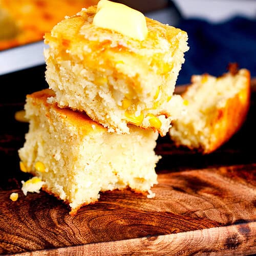 Freshly baked cornbread with frozen corn is stacked on a brown board resting on a blue napkin, with melted butter on the top.