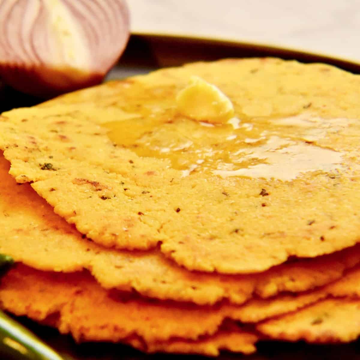 A black plate with cooked Makki ki roti (cornmeal flatbread) topped with a dollop of butter and surrounded by fresh vegetables.