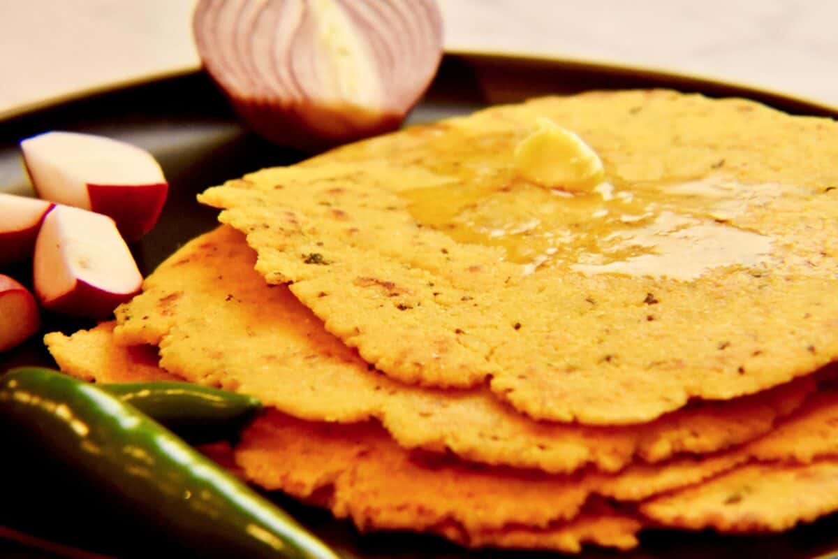 A black plate with cooked Makki ki roti (cornmeal flatbread) topped with a dollop of butter and surrounded by fresh vegetables.