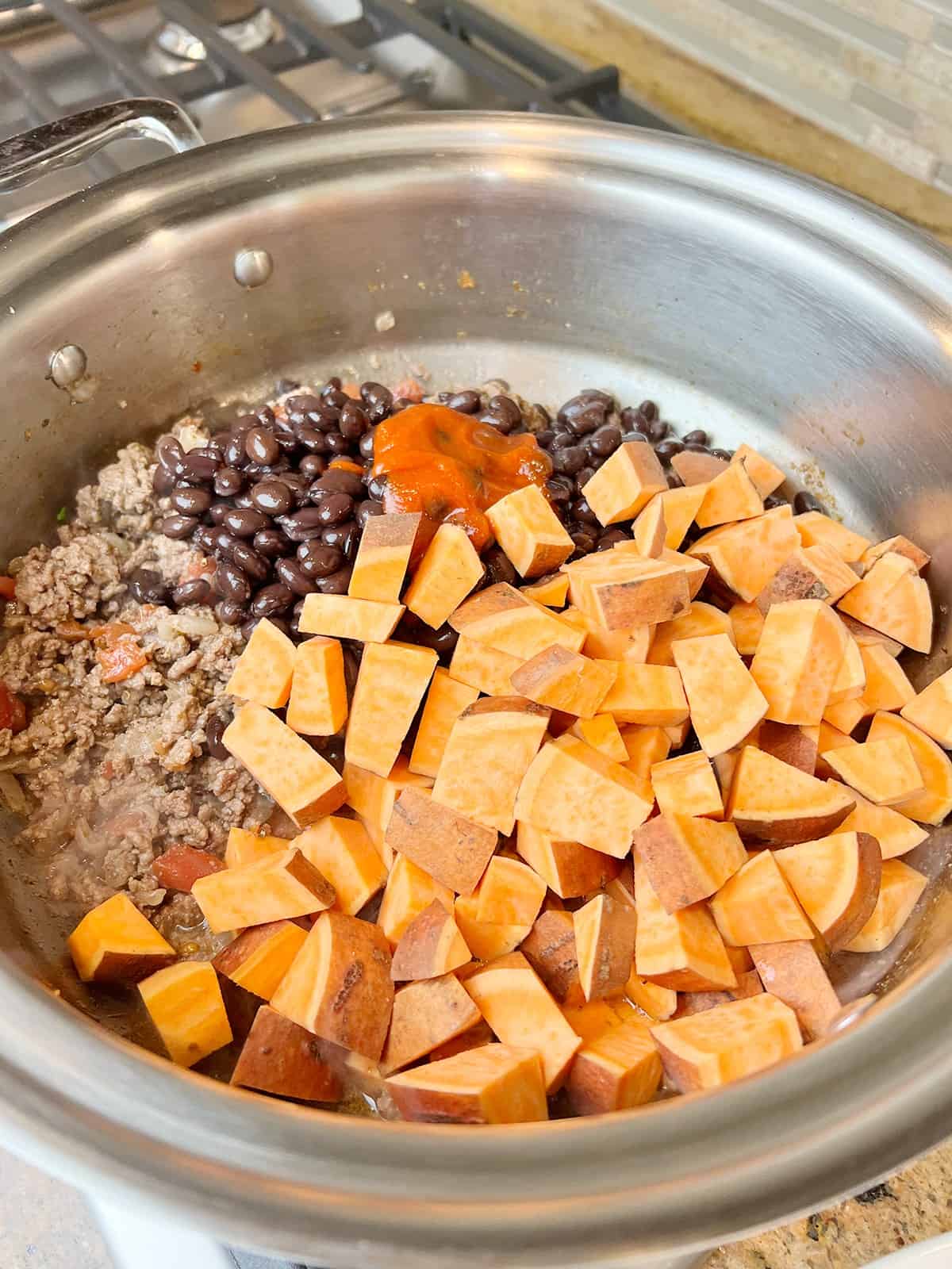 Onions, garlic paste, ground beef, sweet potatoes, black beans and sriracha in a stainless steel pot.