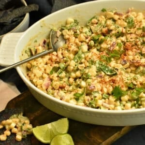 brown board with a white bowl full of corn salad with bue tortilla chips and limes scattered around it.