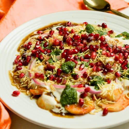 Aloo chaat made with boiled sliced potatoes, yogurt, onions, chutneys and pomegranate seeds is served in an off white plate.