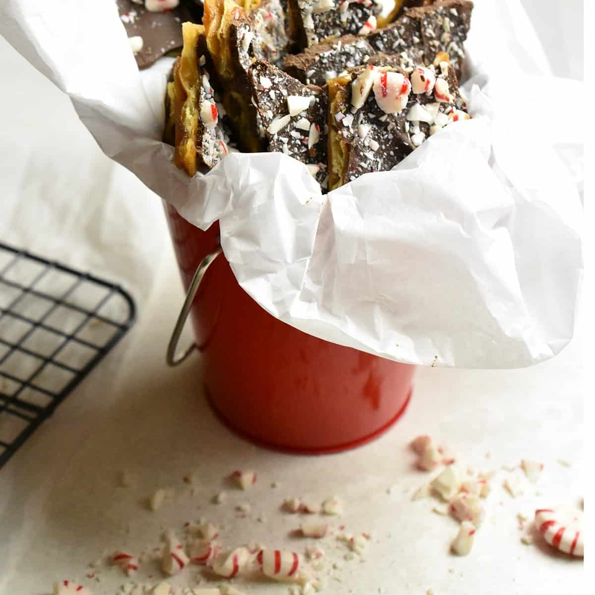 White countertop with a dark metal cooling rack with crack candy resting on a red napkin, crushed peppermint scarred on the counter and a red tin pail lined with white tissue full of the crack candy.