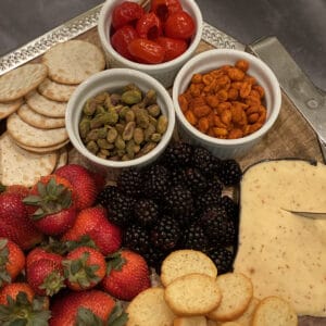 wood tray with silver rim, filled with red and blue berries, marinated red peppers, nuts and crackers.