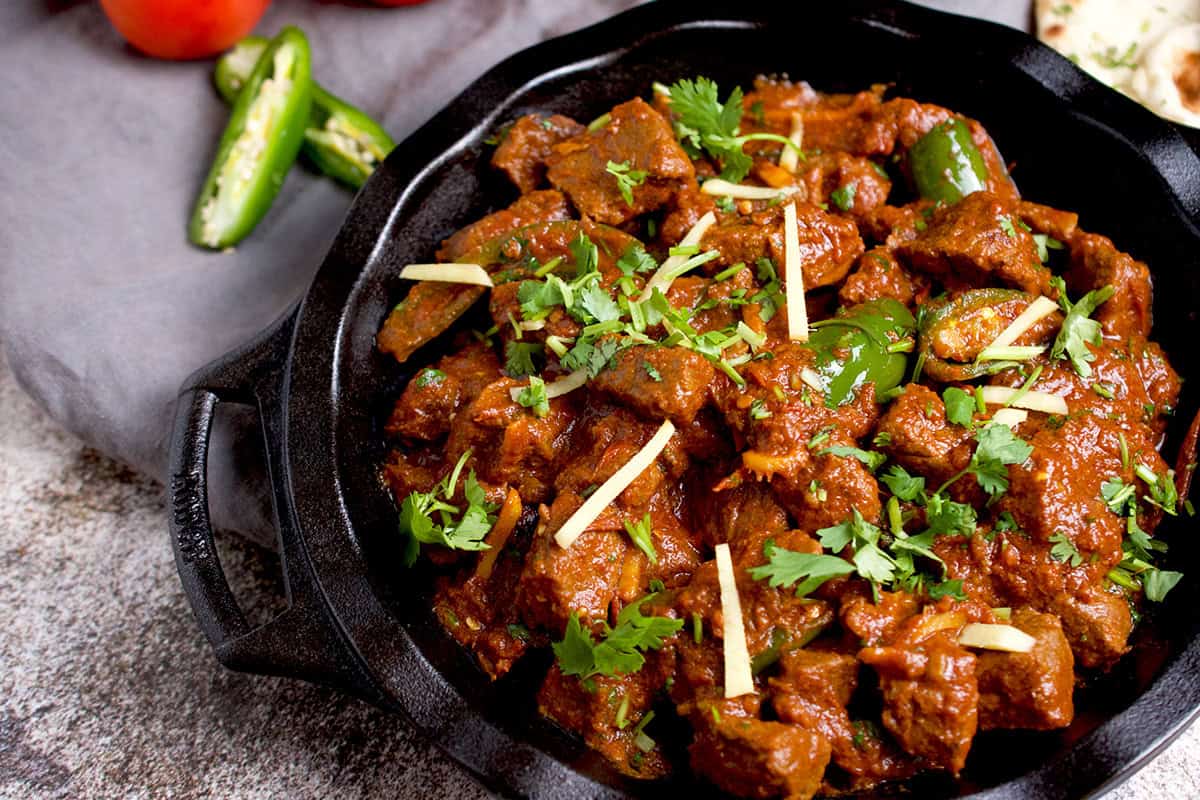 Cooked karahi gosht is in a black skillet with naan in the background.