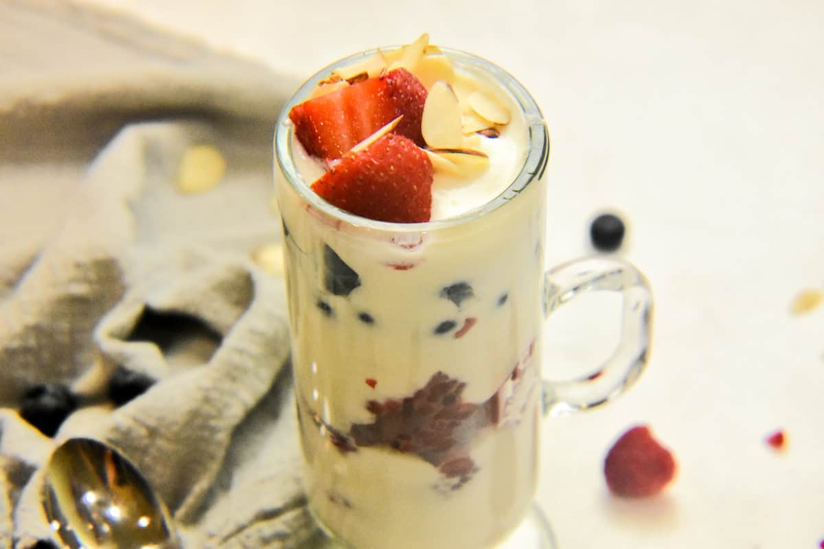 A tall glass filled with whipped ricotta breakfast parfait, layered with mixed berries and topped with a garnish of almonds on a white counter with a grey napkin and scattered berries.