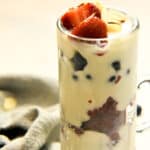 A tall glass filled with whipped ricotta breakfast parfait, layered with mixed berries and topped with a garnish of almonds on a white counter with a grey napkin and scattered berries.