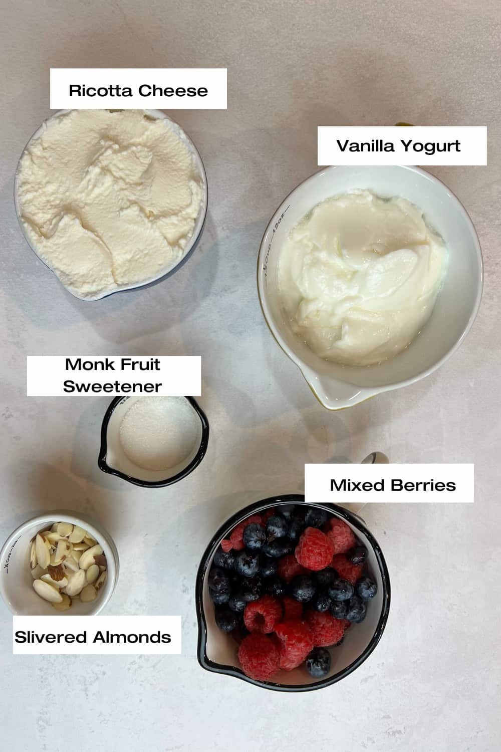 white counter-top with bowls filled with, ricotta cheese, vanilla yogurt, mixed berries, monk fruit sweetener and slivered almonds.