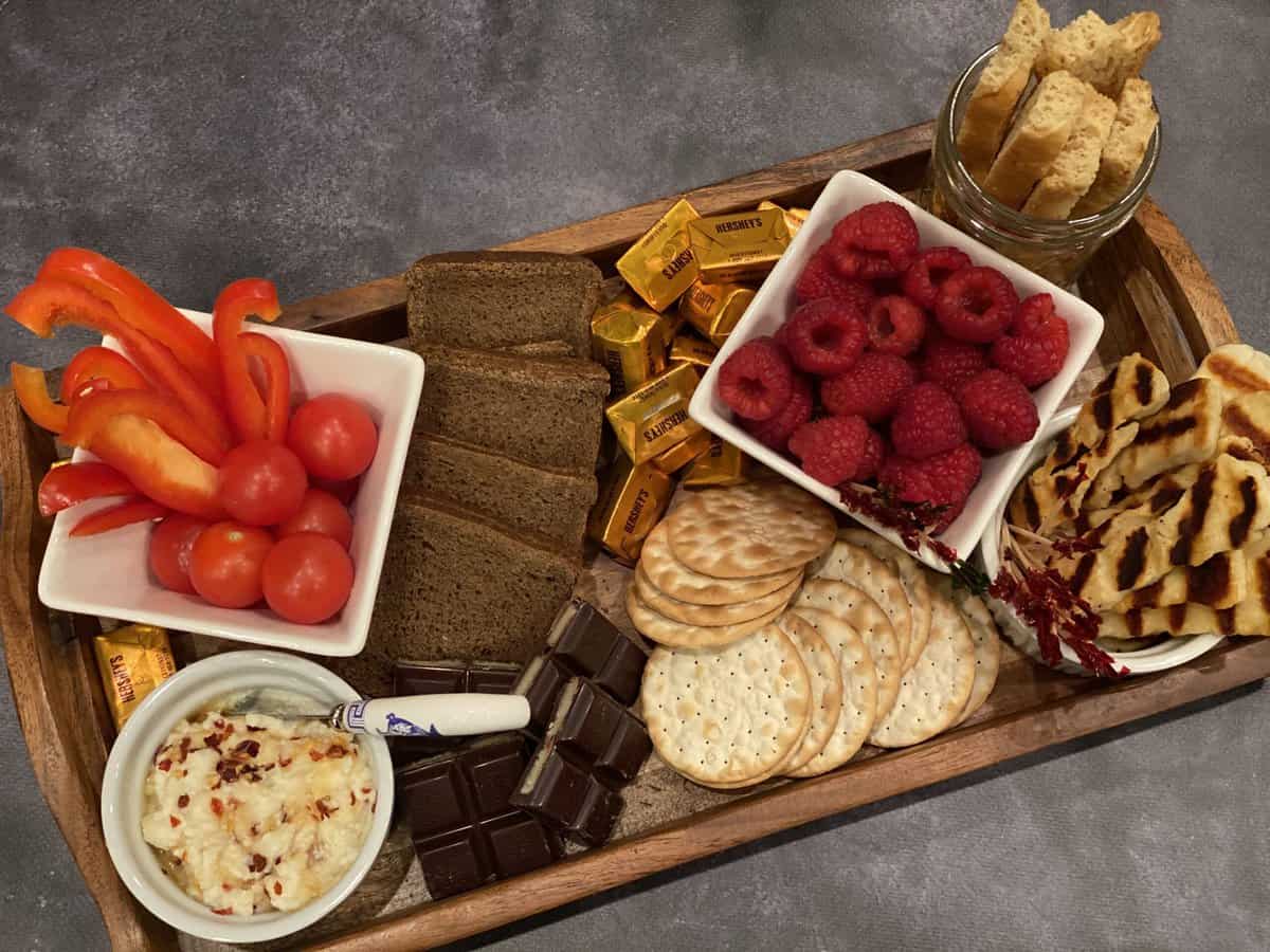 brown tray on a grey background with brown bread, beige crackers, light brown biscotti, white ricotta, white halloumi, brown chocolate, golden wrapped chocolate and red raspberries, red cherry tomatoes and red sliced bell peppers