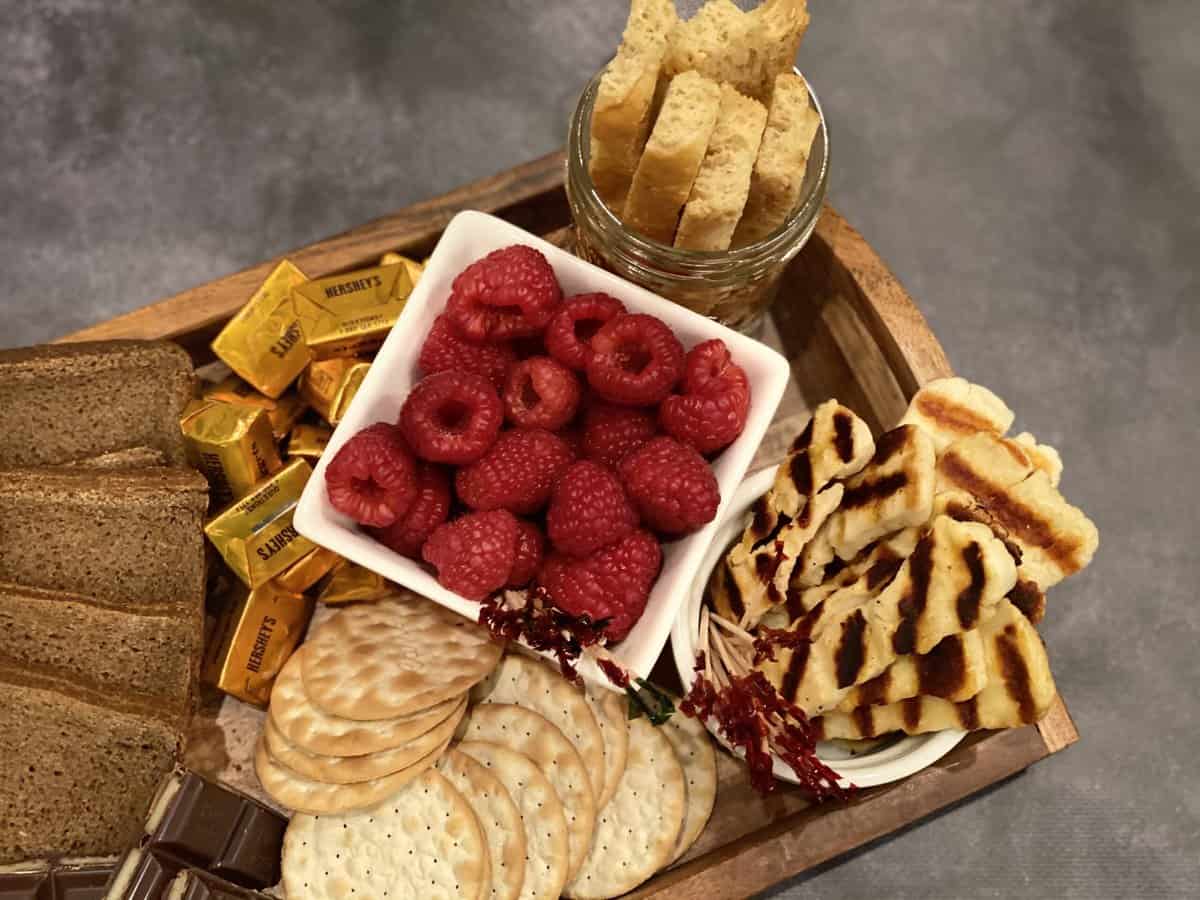 brown tray on greay background with grilled white halloumi, red raspberries, beige crackers, golden wrapped chocolate and savory brown biscotti in a jar