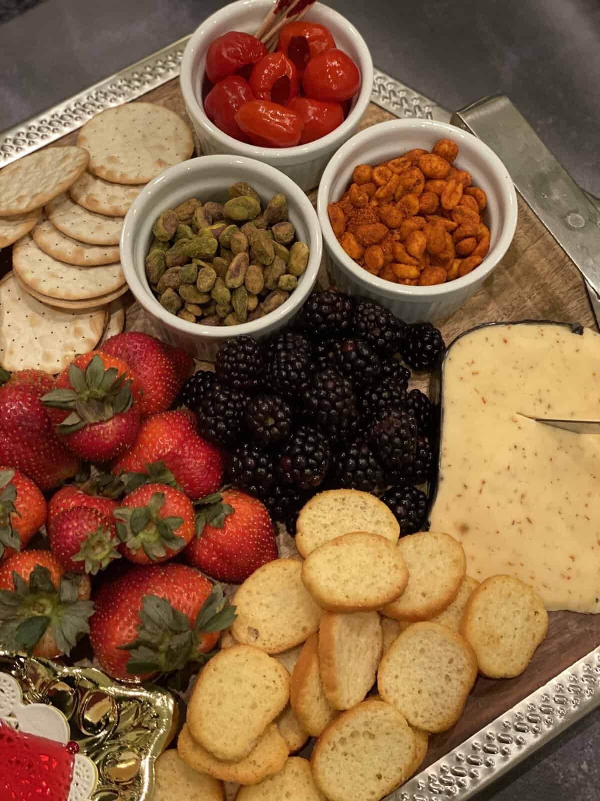brown and silver board with red strawberries, blackberries, cheese with pepper, orange nuts, green nuts, pickled red peppers, two types of crackers