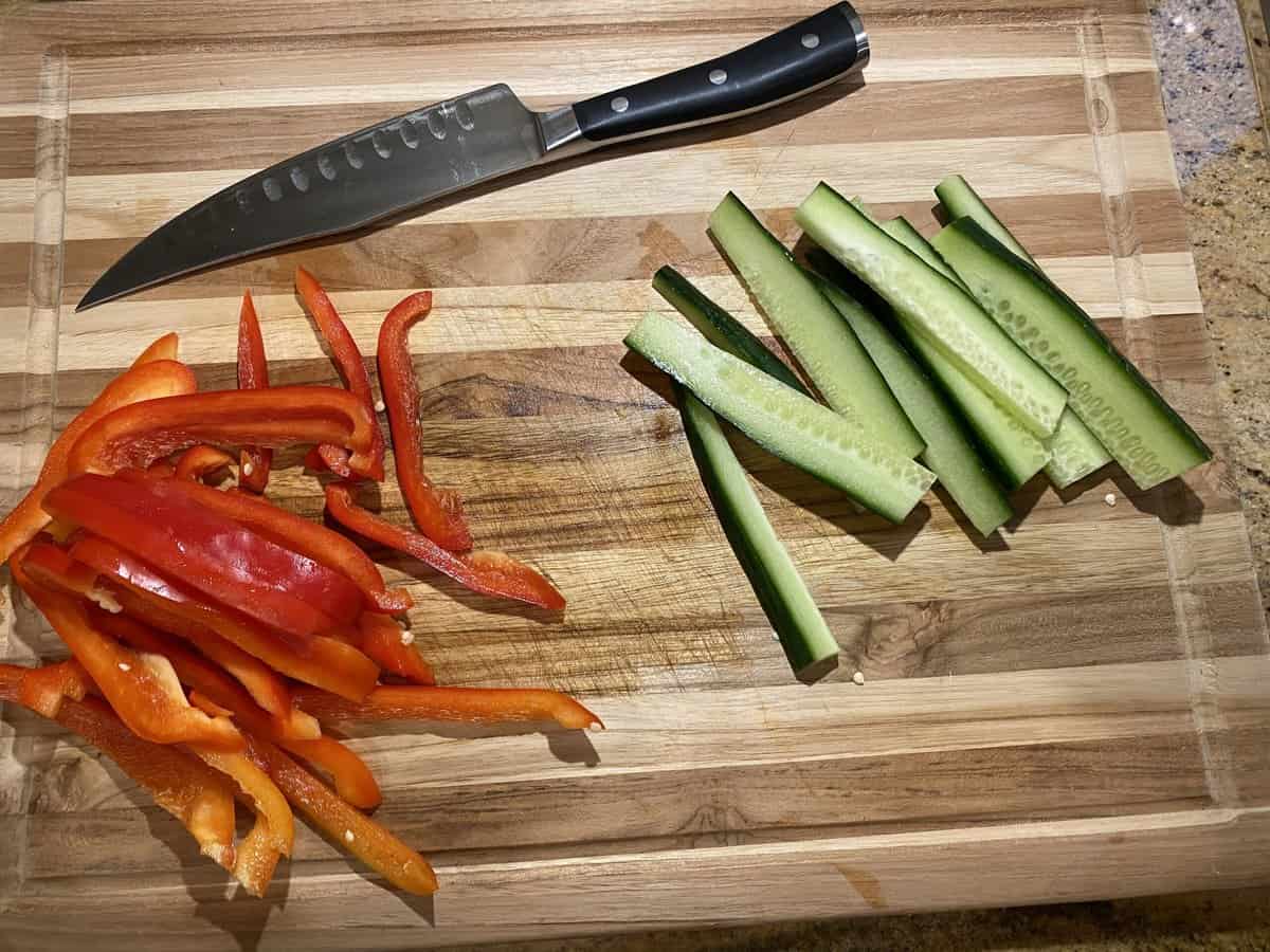 brown wooden chopping board with sliced red bell peppers and cucumbers and a knife alongside them