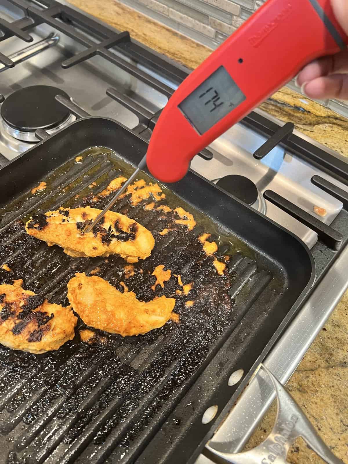 black grill pan on gas stove with cooked chicken tenderloins and red thermometer.