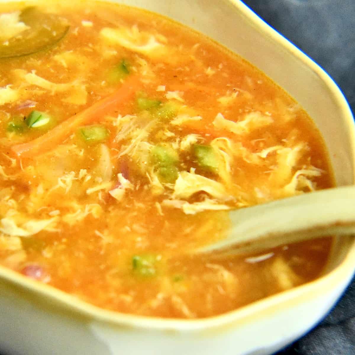 A light grey bowl filled with hot and sour soup in a light orange color with egg ribbons and vegetables floating in it, resting on a blue napkin with condiments in bowls nearby.