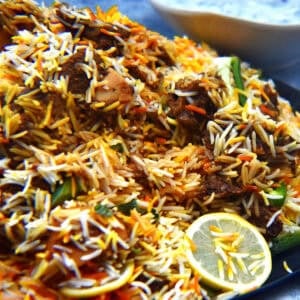 Mutton birayni is plated in a dark metal tray and served with raita and kachumber.