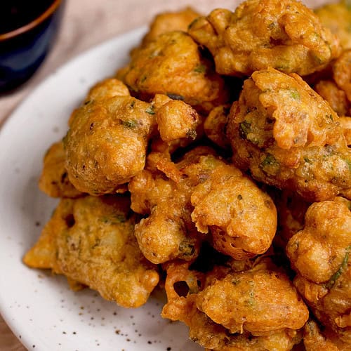 A pakora recipe (with aloo) is plated in a white platter, resting on a light pink napkin with a bowl of chutney in the background.