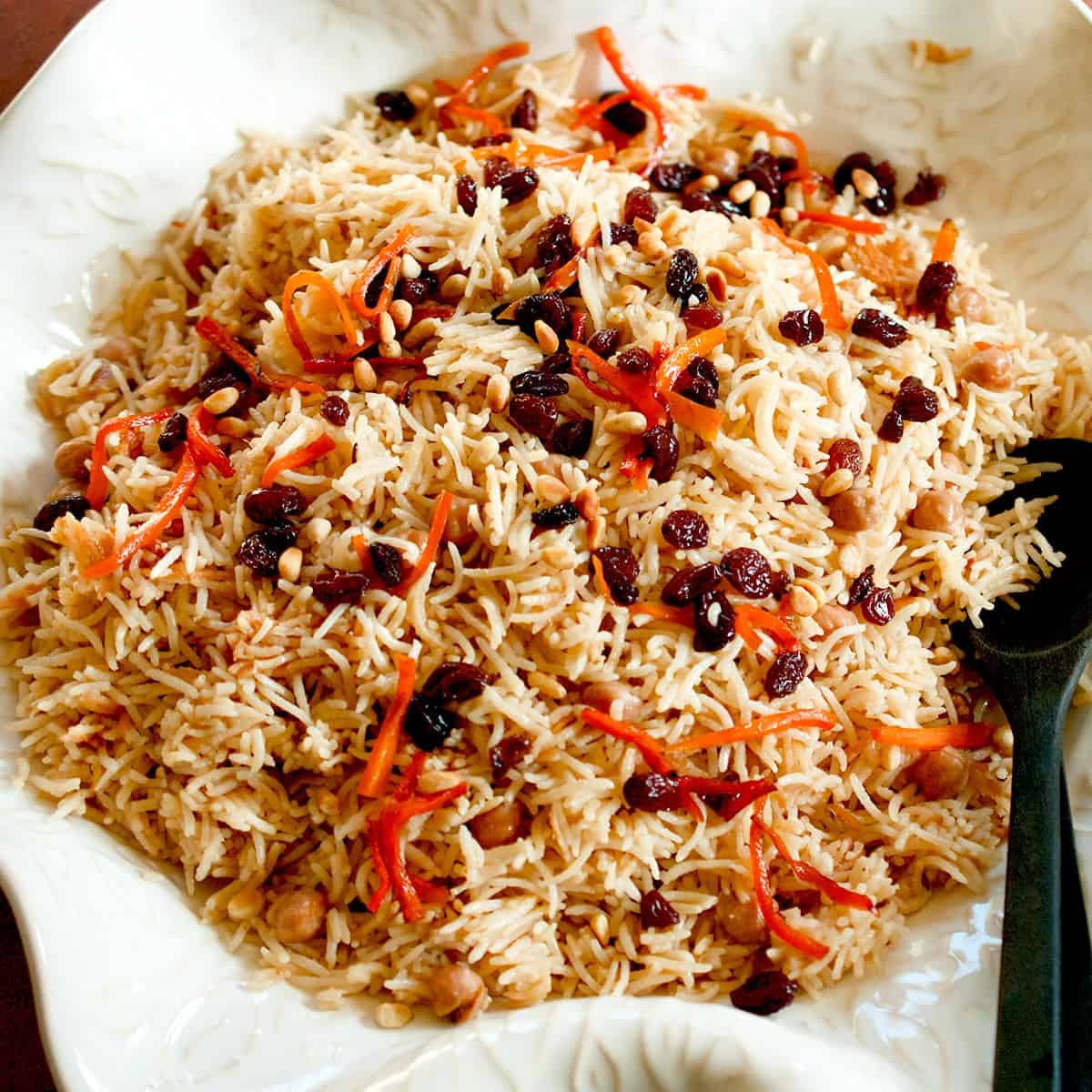 Kabuli pulao (chickpea pilaf), garnished with dark raisins, orange carrots and crunchy pine nuts, is plated in an off-white platter with dark wood spoons.