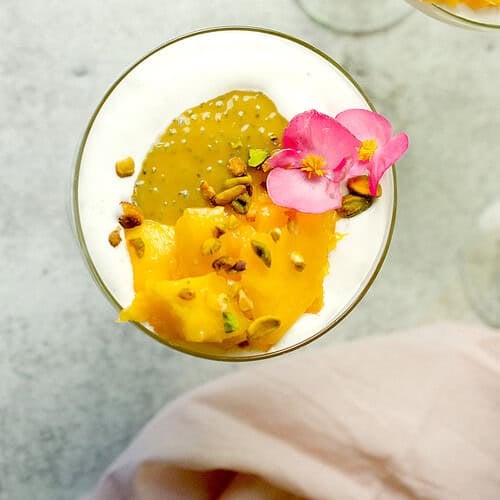 A mango dessert is being served with a garnish of a pink edible flower in a stemmed glass.