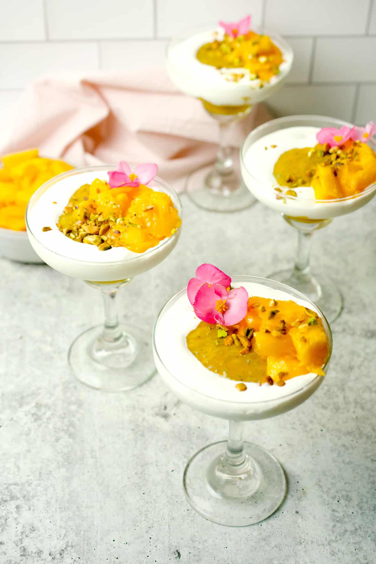An array of stemmed glasses filled with a mango dessert and garnished with a pink flower are ready to be served.