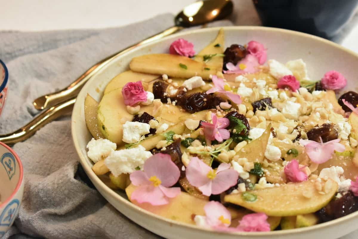 pear salad in a white bowl with dates, goat cheese, hazelnuts, pink flowers and a balsamic glaze.