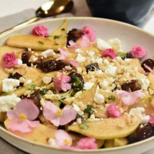 pear salad in a white bowl with dates, goat cheese, hazelnuts, pink flowers and a balsamic glaze.
