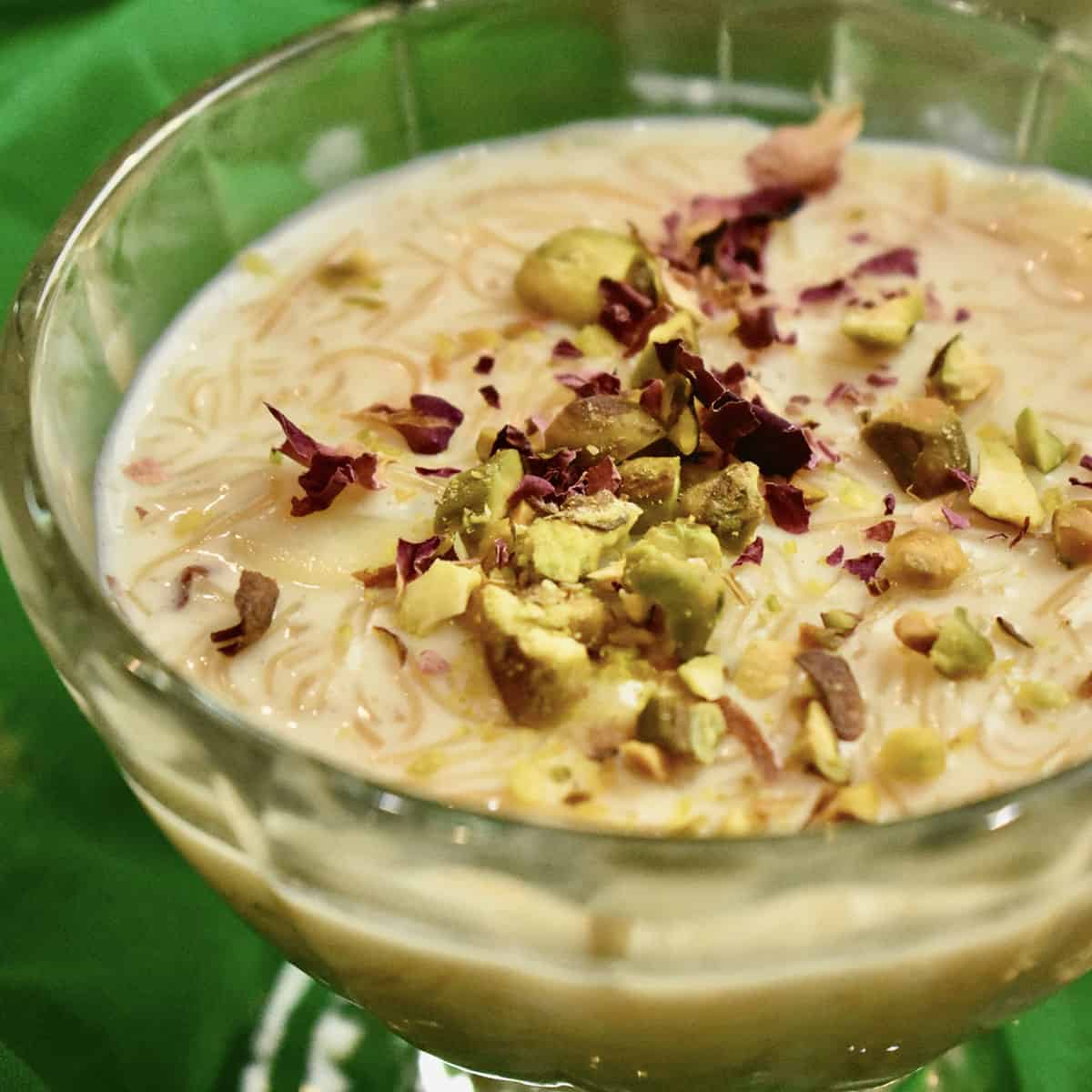 A green gauze scarf and a glass dessert bowl filled with vermicelli kheer topped with pistachios and rose petals.