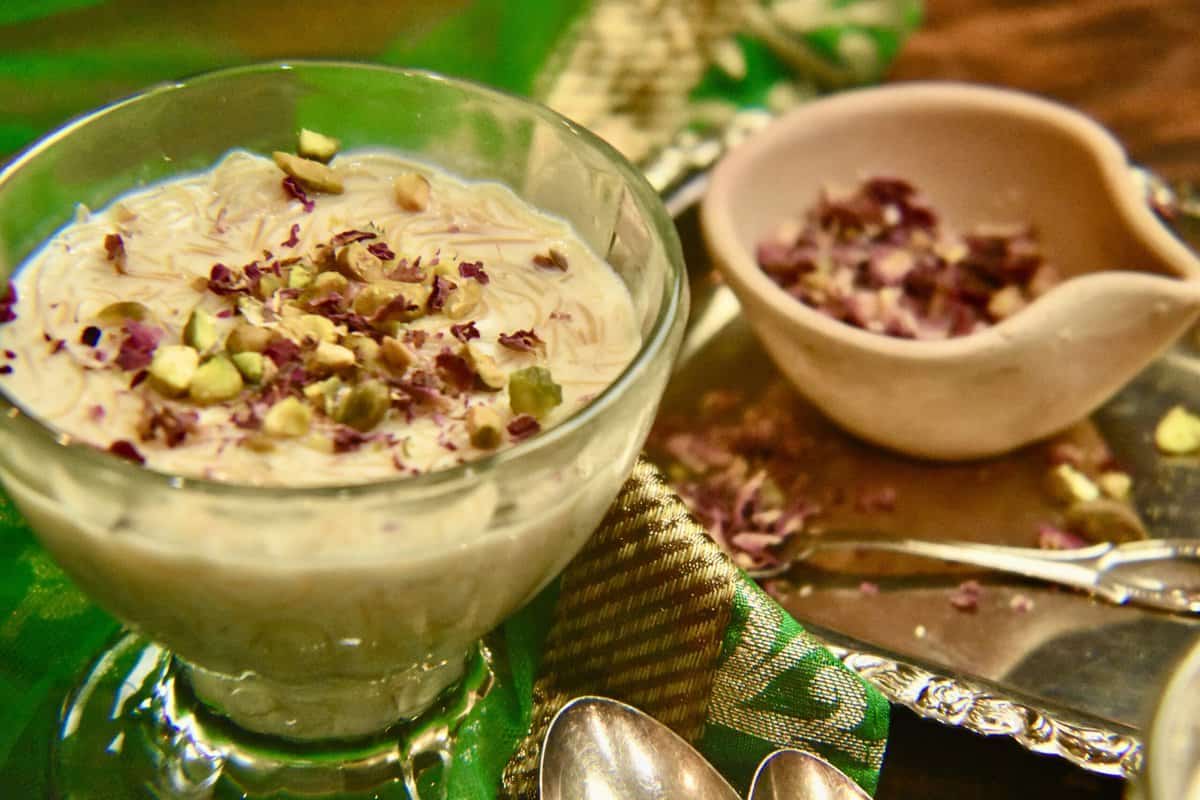 brown table with a green scarf and a dessert bowl of sheer khurma topped with pistachios and rose petals.