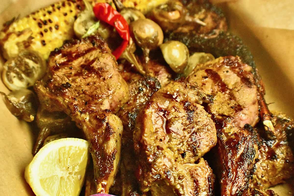 Tandoori grilled lambchops in a tray lined with parchment paper, aerved with grilled corn, roasted baby egg plants, pickled red thai chilies and fresh lemon.