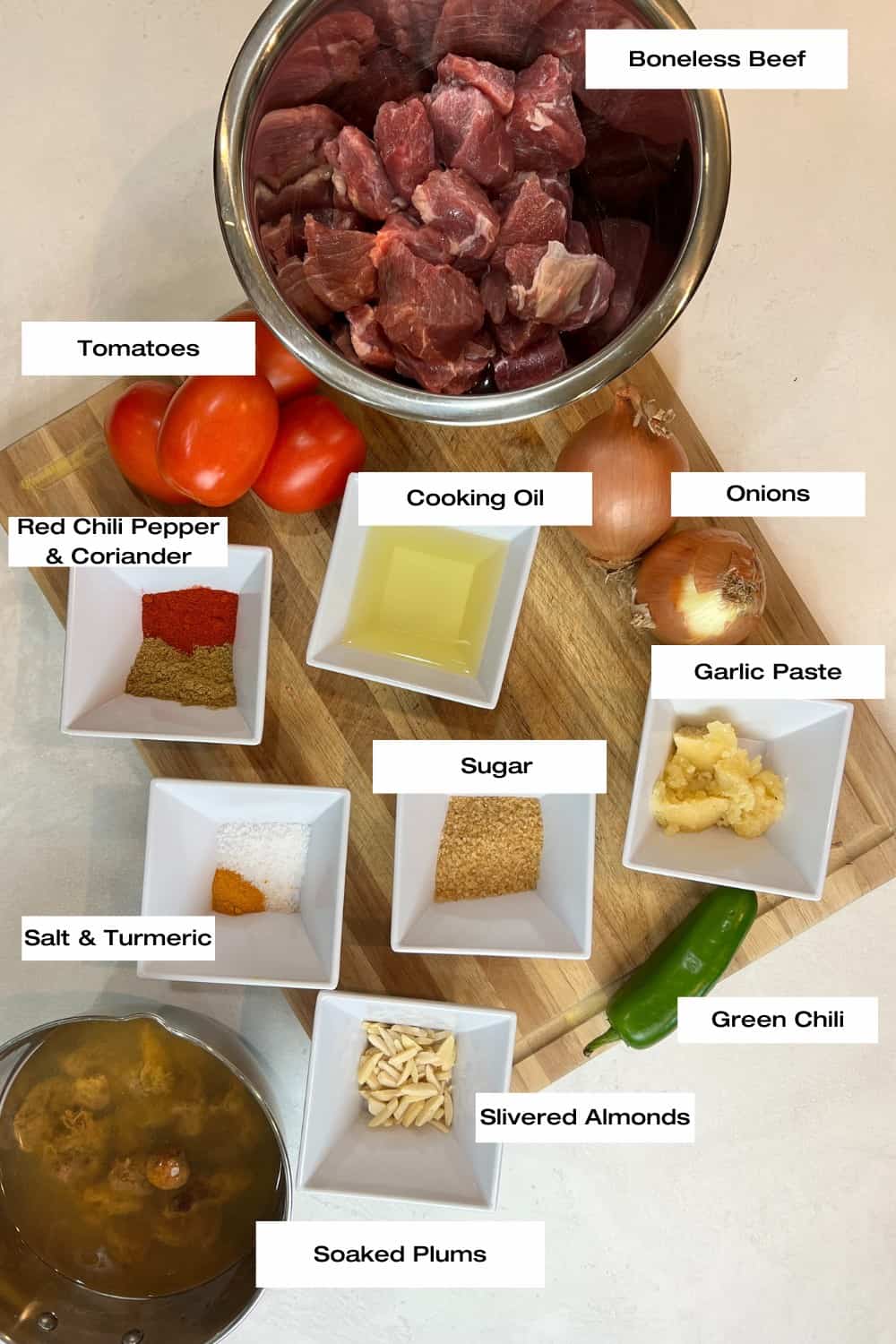 White counter with bowls filled with boneless beef, tomatoes, soaked plums, slivered almonds, sugar, cooking oil, assorted spices, onions, a green chili and garlic paste.